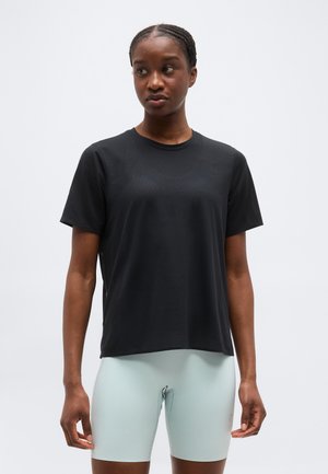 Femme aux cheveux tressés portant un haut de sport noir à manches courtes et un short de cyclisme ajusté gris clair, sur un fond uni.