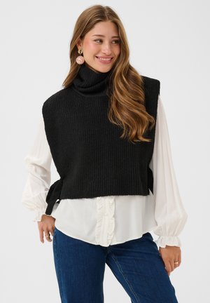 Jeune femme portant un pull noir sans manches à col roulé par-dessus une blouse blanche et un jean bleu, souriante et regardant vers sa gauche.