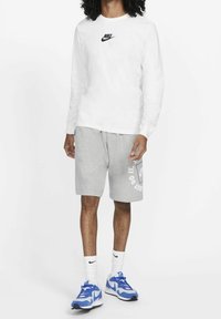 Camisa blanca de manga larga con el logo negro de Nike, pantalones cortos grises con el texto "Just Do It", calcetines blancos y zapatillas deportivas azules con acentos negros.