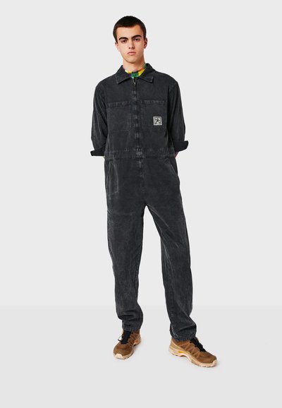 Schwarzer Jeans-Overall mit langen Ärmeln, einem Frontreißverschluss und einer aufgesetzten Tasche. Verfügt über sichtbare Nähte und eine entspannte Passform. Braune Sneakers getragen.
