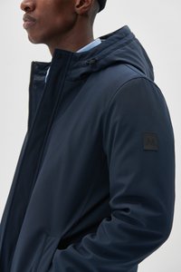 Chaqueta azul marino con capucha, hecha de material suave. Presenta un parche de logo negro en la manga y un cierre frontal con cremallera.