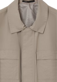 Beige leichte Jacke mit einem umlegbaren Kragen, knopflosem Frontbereich und zwei aufgesetzten Taschen. Glatte Textur mit sichtbaren Nähten.