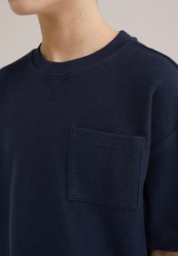 Bordo modra pletena majica s kratkimi rokavi, ki ima okroglo izrez in žep na levi strani ter teksturirano tkanino.