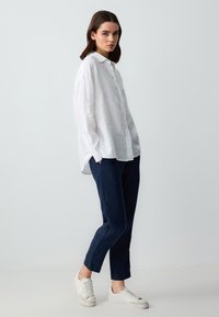 Chemise blanche en lin à boutons avec des manches longues, associée à un pantalon navy longueur cheville et des baskets blanches. Design simple et décontracté.