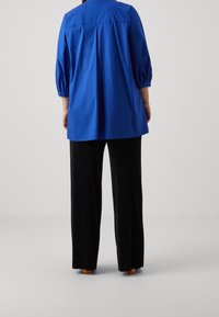 Blå, löst sittande blus med 3/4 ärmar, knappmanchetter och rund halsringning, i kombination med svarta, vida ribbade byxor.