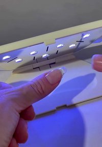 Tragbare Nagellampe mit weißem Gehäuse, LED-Lichtern und reflektierender Oberfläche. Die Finger zeigen ein klares Nageldesign unter blauem Licht.