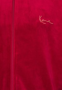 Veste zippée en velours rouge avec un subtil logo doré sur la poitrine. Le tissu a une texture lisse et douce avec une finition brillante.