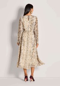 Langes, schlangenmusteriges Kleid in Beigetönen und Brauntönen mit langen Ärmeln, tailliert und mit Detail im Schlüsselrücken, kombiniert mit burgunderfarbenen High Heels.