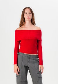 Pull côtelé rouge décolleté avec de longues manches, design ajusté et encolure large, associé à un pantalon cargo gris.