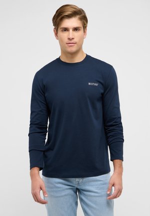 Junger Mann, der ein marineblaues Langarmshirt mit dem "MUSTANG"-Logo auf der Brust und hellblaue Jeans trägt, steht vor einem schlichten weißen Hintergrund.