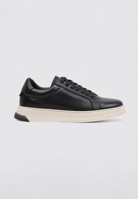 Zapatilla negra de cuero con punta redonda, diseño plano con cordones y suela de color crema contrastante con un pequeño tacón. Textura lisa.