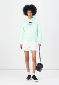 Licht mintgroene hoodie met een frontlogo, gecombineerd met witte shorts. Het model draagt zwarte sneakers en houdt een zwarte, ronde tas vast.