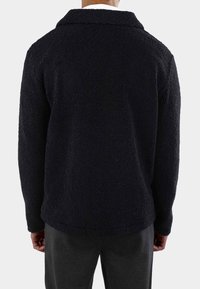 Sort fleece jakke med en blød tekstur, krave-design og drop-skulder. Har en afslappet pasform og afrundet kant, vist fra bagsiden.