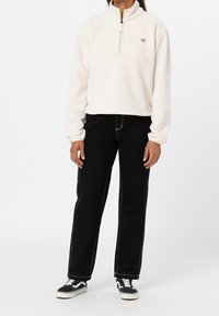 Pullover in pile color crema con mezza zip, caratterizzato da un piccolo logo e polsini elasticizzati. Abbinato a pantaloni neri e scarpe da ginnastica nere.