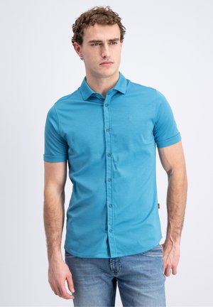 Ung mand iført en tætsiddende kortærmet blå skjorte med knapper og lyseblå jeans, stående foran en ensfarvet baggrund.