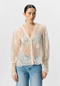 Blonde skjorte med V-udskæring, cremefarve, gennemskinneligt stof, puffede ærmer, blomster Mønstre og knaplukning foran. Sammen med lyseblå jeans.