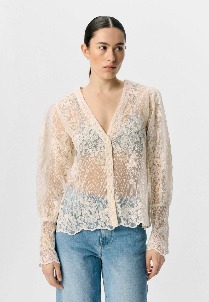Blonde skjorte med V-udskæring, cremefarve, gennemskinneligt stof, puffede ærmer, blomster Mønstre og knaplukning foran. Sammen med lyseblå jeans.