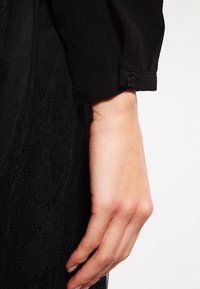 Blusa nera a maniche lunghe in tessuto a trama, con polsino abbottonato e dettaglio in pizzo visibile sotto. Ingrandimento di una mano che tiene la manica.