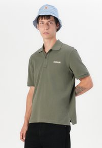 Oliven grønn polo skjorte med korte ermer, med en subtil logo på brystet. Modellen har på seg en lys blå bøttehatt.
