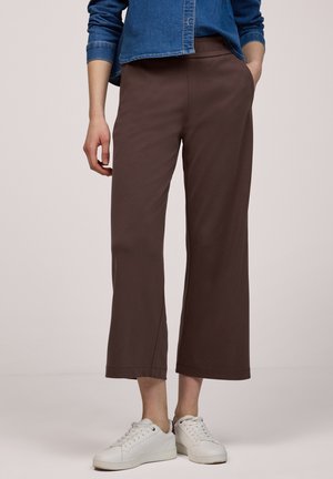 Persona con pantalones marrones anchos y cortos, zapatillas blancas y una camisa de mezclilla azul, con una mano en el bolsillo.