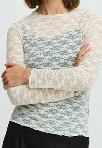 Haut en dentelle avec un design crème et bleu clair, manches longues, col rond et un look superposé avec des motifs floraux.