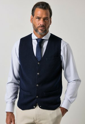 Mann mit Bart, der ein weiß gestreiftes Hemd, eine marineblaue Weste, eine marineblaue Krawatte und beige Hose trägt, steht mit einer Hand in der Tasche.