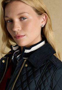 Geacă matlasată bleu marin cu accesorii aurii, având un guler înalt cu dungi albe și bleu marin dedesubt. Cercei cu picior de argint vizibili.