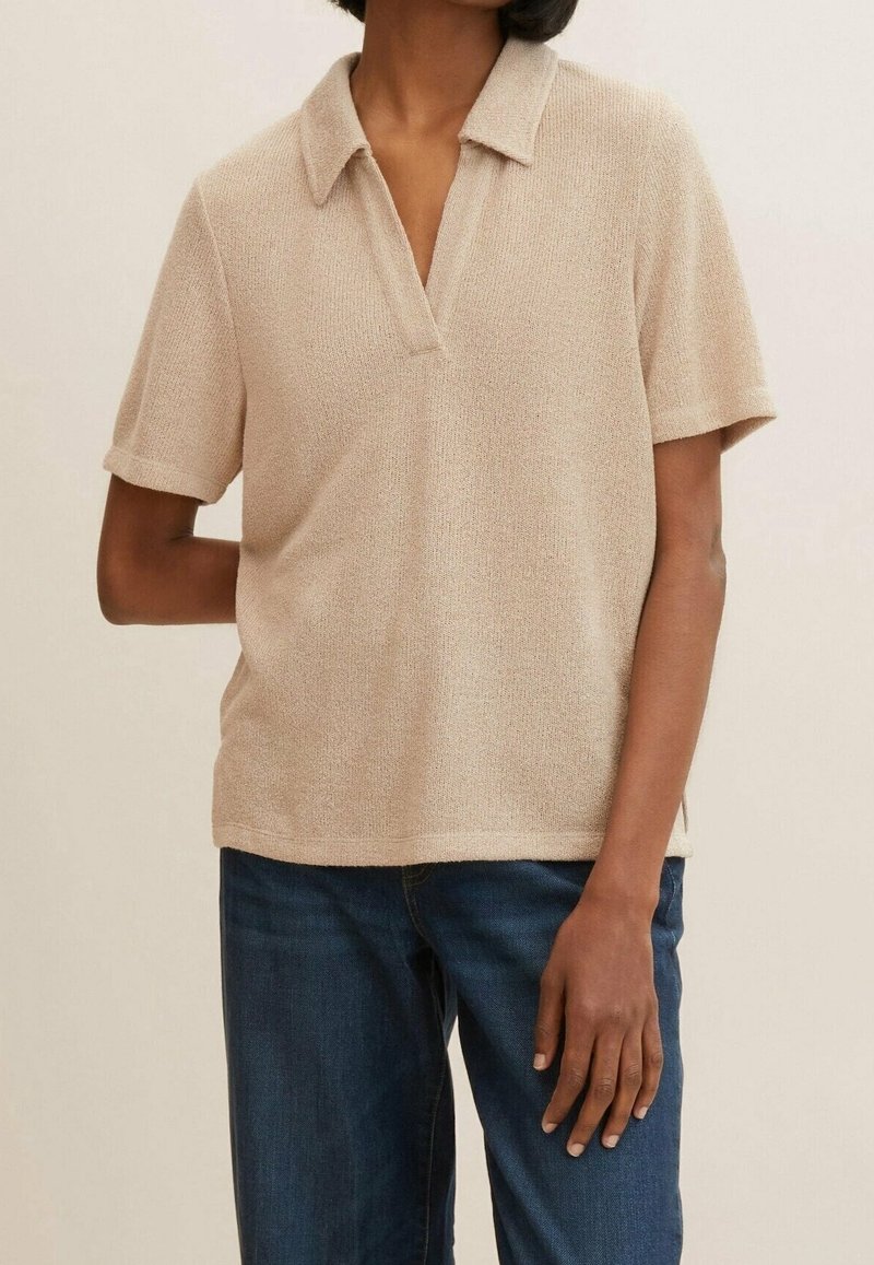 Beige polo tričko s krátkým rukávem a texturovaným úpletem, se mělčím výstřihem ve tvaru V, nošené přes tmavě modré džíny.