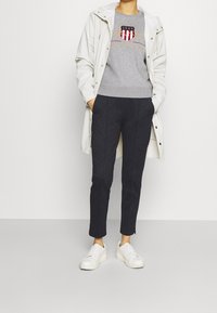 Grå sweatshirt med et logo, hvid vandtæt frakke, mørke bukser og hvide sneakers. Frakken har trykknapper og en hætte.