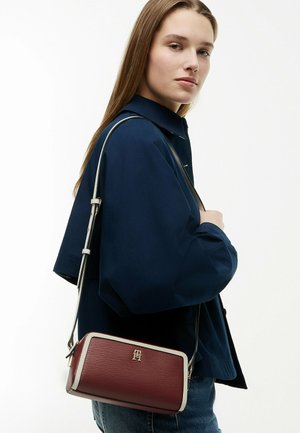 Mujer con el pelo largo, llevando una chaqueta azul marino y vaqueros, y que lleva un bolso rectangular burdeos con bordes claros y una correa para el hombro.