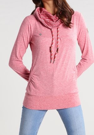 Vrouw draagt een roze trui met lange mouwen, een patroon col en koordjes, gecombineerd met lichtblauwe spijkerbroek.