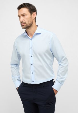 Hellblaues langärmliges Hemd mit Button-Down-Kragen, ausgestattet mit schwarzen Knöpfen und einer feinen Textur. Kombiniert mit dunklen Hosen.
