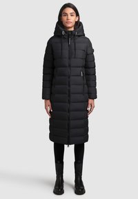 Manteau rembourré noir avec capuche, présentant un design matelassé, une fermeture éclair sur le devant, des poches latérales et une coupe mi-mollet. Porté par-dessus un pantalon noir et des bottes.