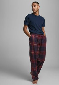Camiseta de manga corta azul marino combinada con pantalones de cuadros burdeos holgados con líneas amarillas. El modelo está descalzo, con las manos en los bolsillos.
