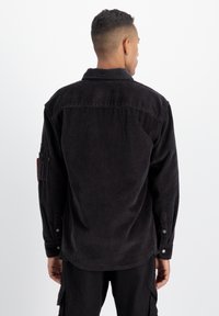 Camicia in costina nera con maniche lunghe, polsini con bottoni e yoke sulla schiena. Presenta una tasca a toppa sul braccio sinistro e un tessuto testurizzato.