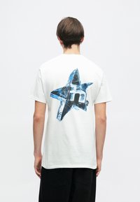 Άσπρη βαμβακερή μπλούζα με κοντό μανίκι που διαθέτει μια μπλε και μαύρη γραφική παράσταση αστεριού στο πίσω μέρος, με ένα μοντέρνο λογότυπο στο κέντρο. Χαλαρή εφαρμογή.