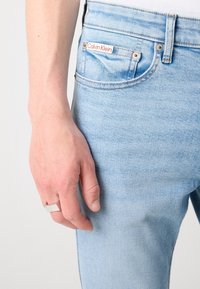 Jeans de mezclilla azul claro con un ajuste casual, que presentan una etiqueta roja de Calvin Klein en el bolsillo, un desvanecimiento sutil y detalles de costura estándar.