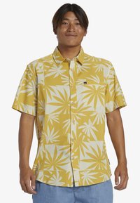 Camicia a maniche corte di colore giallo con motivo a foglia di palma bianca, realizzata in tessuto leggero. Design con bottoni e una piccola etichetta con logo.