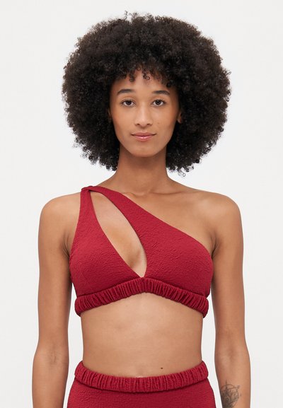Asymetrický červený bikini top s texturovanou tkaninou, který má výřezový design a nařasenou pásku pod prsy. Celkově hladký povrch.