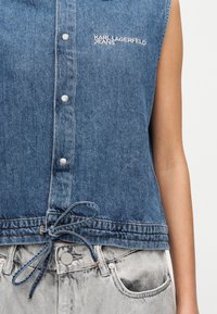 Μπλε τζιν γιλέκο χωρίς μανίκια με κουμπιά τύπου snap και κορδόνι δεμένο στη μέση, με το λογότυπο "Karl Lagerfeld Jeans" πάνω από το αριστερό στήθος.