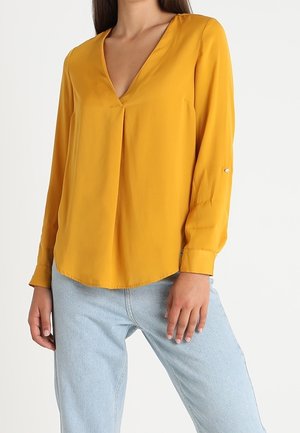 Femme portant un chemisier jaune moutarde à manches longues et col en V associé à un jean en denim bleu clair sur fond blanc.