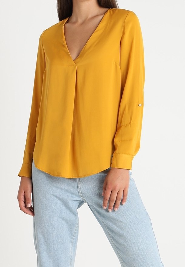 Femme portant un chemisier jaune moutarde à manches longues et col en V associé à un jean en denim bleu clair sur fond blanc.