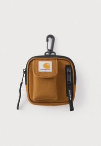 Καφέ υφασμάτινη θήκη Carhartt με εμπρόσθια τσέπη, μαύρο φερμουάρ και μαύρο κλιπ σε απλό λευκό φόντο.