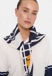 Navy and white patterned scarf with a grid design, featuring a round orange detail. Fabric has a soft texture, draped around the neck.