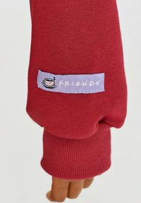 Červená mikina s žebrovanými manžetami, na níž je fialová textilní nášivka s nápisem "F.R.I.E.N.D.S" a grafikou šálku kávy.