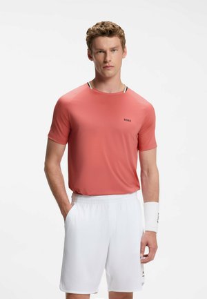 Junger Mann trägt ein korallenfarbenes Kurzarmhemd mit "BOSS"-Logo, weiße Sportshorts und ein weißes Armband, steht mit einer Hand in der Tasche.
