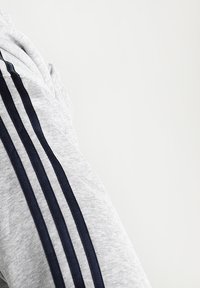 Tejido de sudadera deportiva gris con tres franjas horizontales en azul marino en la manga. El material parece suave y tiene una textura ligera.