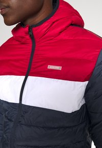 Rød, hvid og marineblå stribet puffy jakke med lynlås, hætte og et logo-applikation. Materialet virker glat og struktureret.