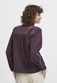 Short-sleeved shirt in deep purple, featuring a smooth texture, button accents, and a relaxed fit with a pleated back design.