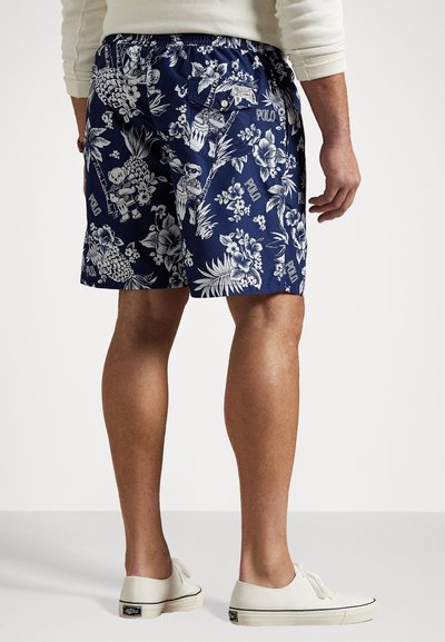 Jūras zila peldbikses ar baltām ziedu un tropu rakstiem; elastīga josta; viena aizmugures kabata ar pogu; apvienotas ar baltām apaviem.